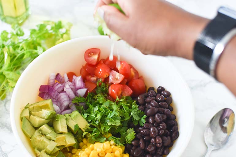 Avocado Black Bean Corn Salad in large bowl. Brown hand squeezing lime overtop